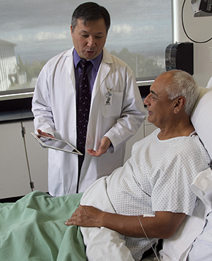 Un proveedor de atención médica sostiene una tableta y habla con un hombre en la cama del hospital.
