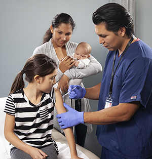 Proveedor de atención médica aplicándole una inyección en el brazo a una niña mientras una mujer con un bebé en brazos mira.